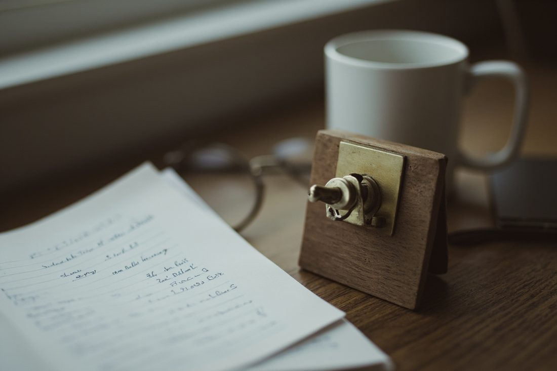 A small brass toggle switch mounted on an oak block on a writer's desk, beside an open notebook of handwritten notes, reading glasses and a white mug in soft window light.