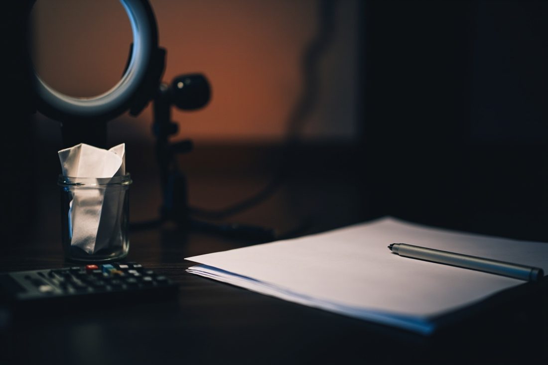 A dark home streaming desk with a ring light and small camera behind a glass jar of folded white paper, a calculator, and a stack of blank paper with a silver pen.
