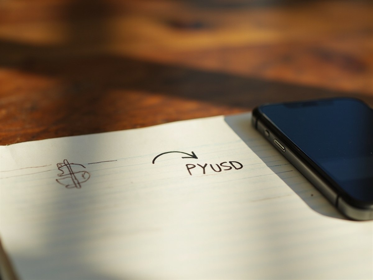 A close-up of a smartphone screen showing a payment balance, beside notepad with a hand-drawn arrow from a dollar sign to a coin labelled PYUSD.