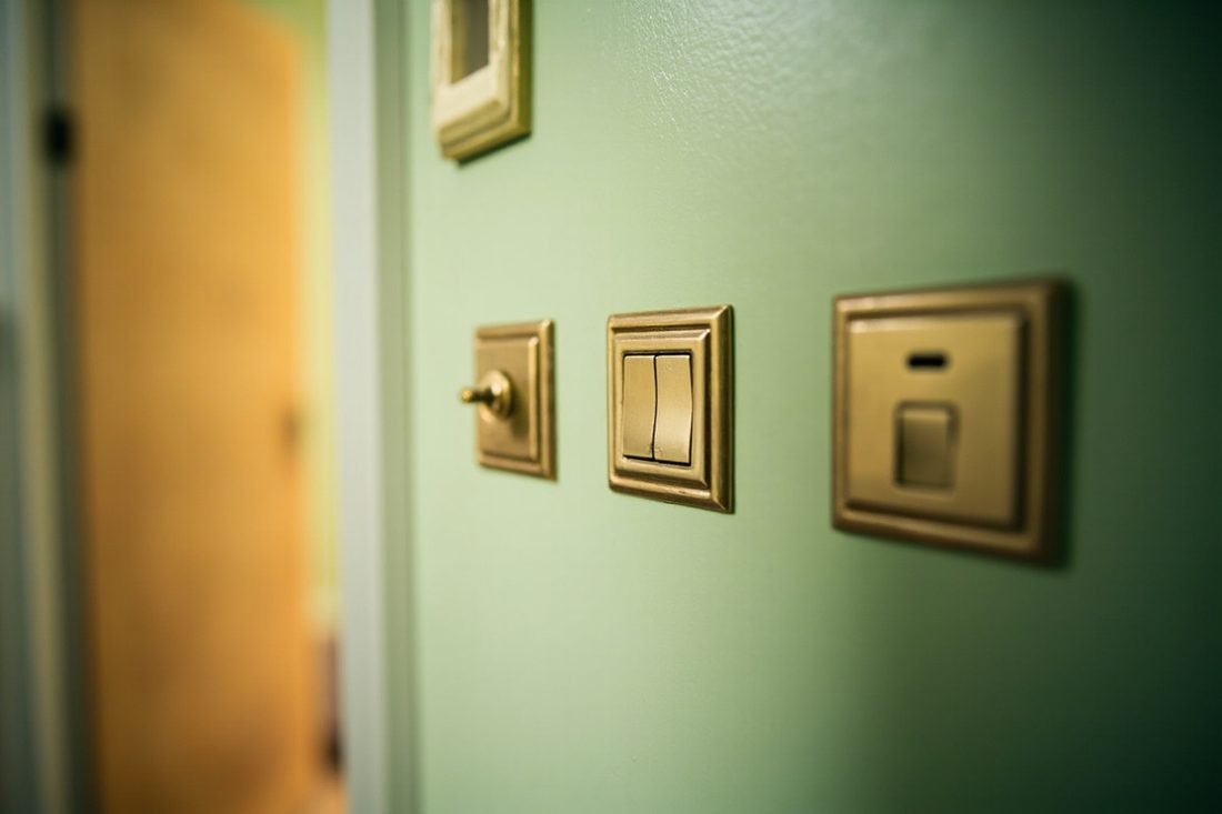 Three brass light switches of different styles on a pale green wall, next to a door left slightly open.
