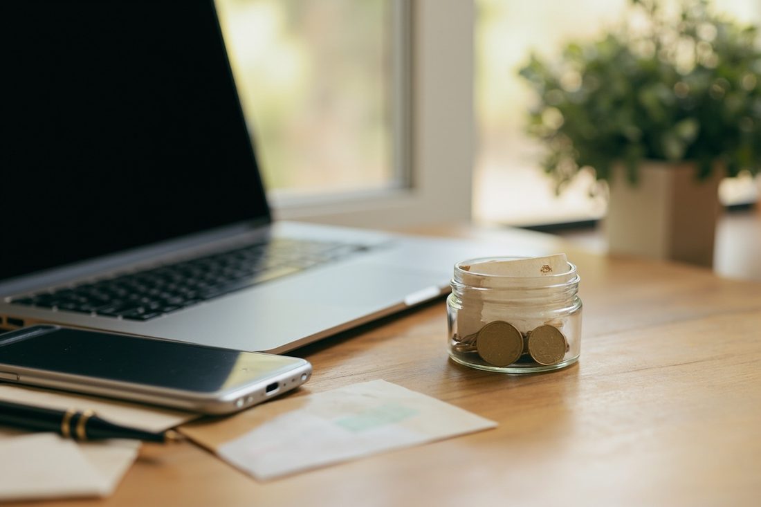 A laptop with a dark screen and a smartphone on a wooden desk by a sunny window, beside a small glass jar holding folded paper notes and a few plain brass coins, a paper envelope and a black pen.
